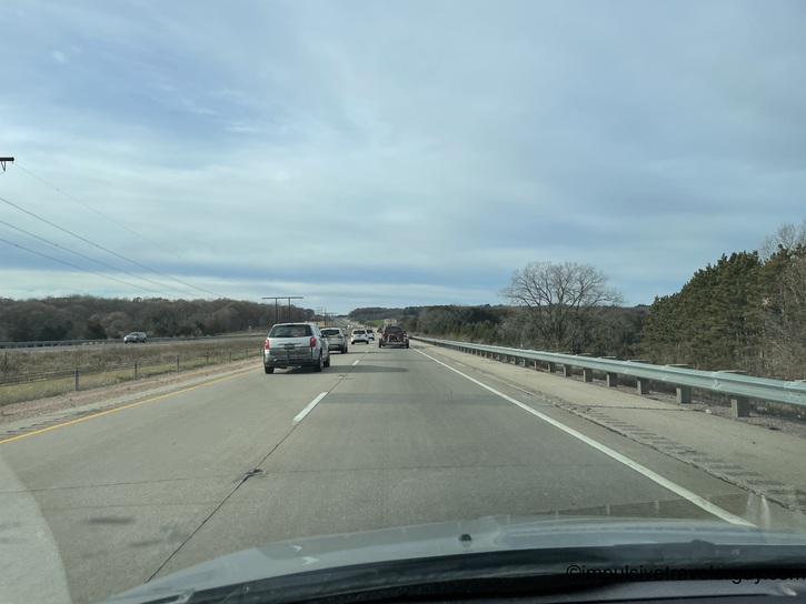 Multi-lane highway driving scene with vehicles ahead, guardrail visible on right side, bare trees and overcast sky