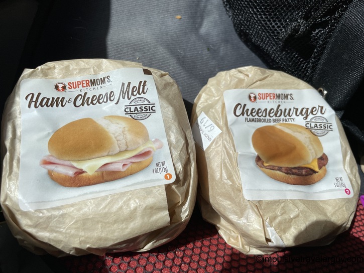 Two SuperMom's frozen sandwich packages (Ham & Cheese Melt and Cheeseburger) in their brown paper packaging resting on a dark fabric car seat
