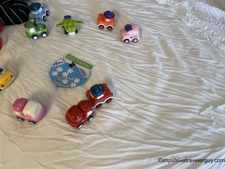 Hotel bed with colorful toy vehicles scattered on white comforter, including dump trucks, cars in various colors, and Christmas-themed decorative item