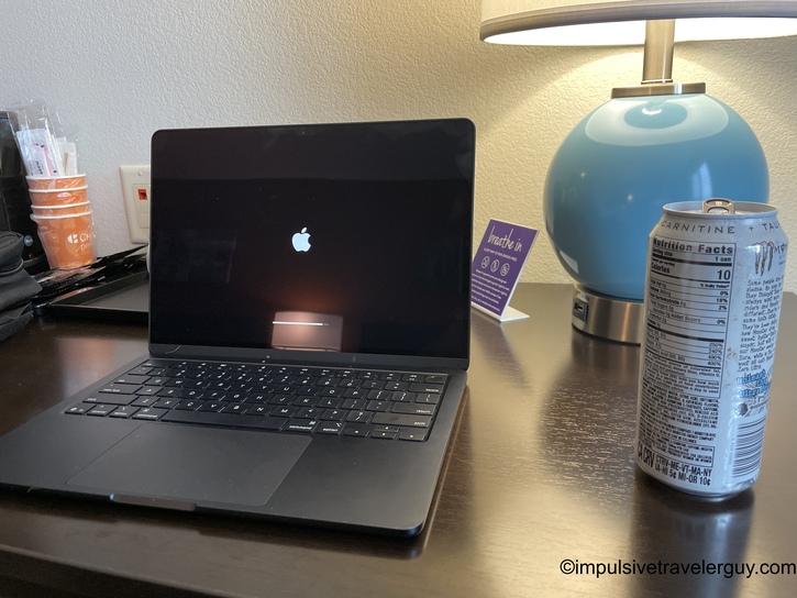 MacBook laptop on hotel desk showing Apple logo and loading bar on screen, with Monster energy drink, lamp, and coffee supplies nearby