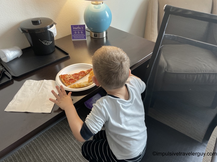 Young child in white shirt sitting at hotel desk eating pizza slice with breadstick on white plate