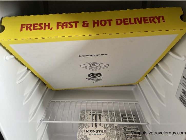 Interior of mini refrigerator showing Monster energy drinks stored on bottom shelf with yellow corrugated pizza box holder on top reading Fresh, Fast & Hot Delivery!