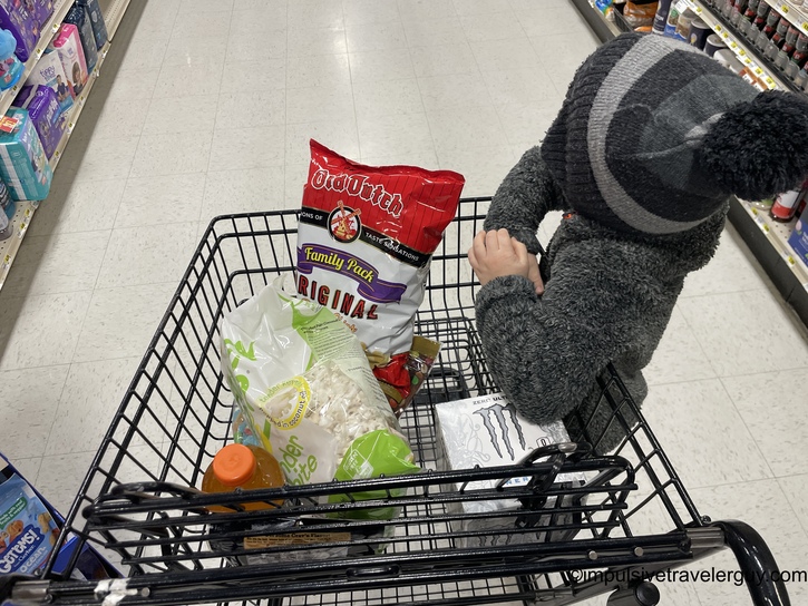 Overhead view of a shopping cart containing groceries including a bag of Crunch chips, Asian noodles, Monster energy drink, and other snack items, with a child's hand visible on the cart handle