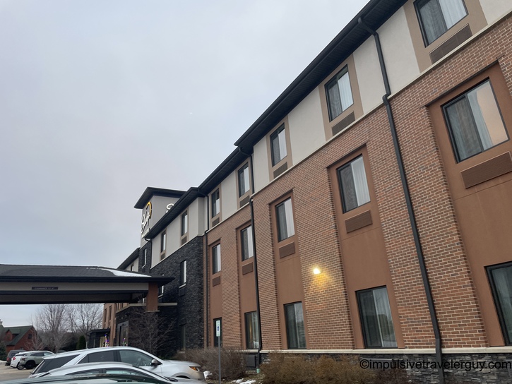 Exterior view of modern Sleep Inn hotel building with tan brick and beige facade, multiple stories, covered entrance portico, and parking lot in overcast conditions