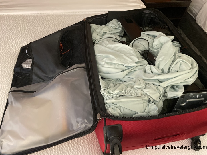 Open red wheeled suitcase on hotel bed containing neatly packed light-colored clothing, bedsheets, cables, and packing cubes with mesh compartment