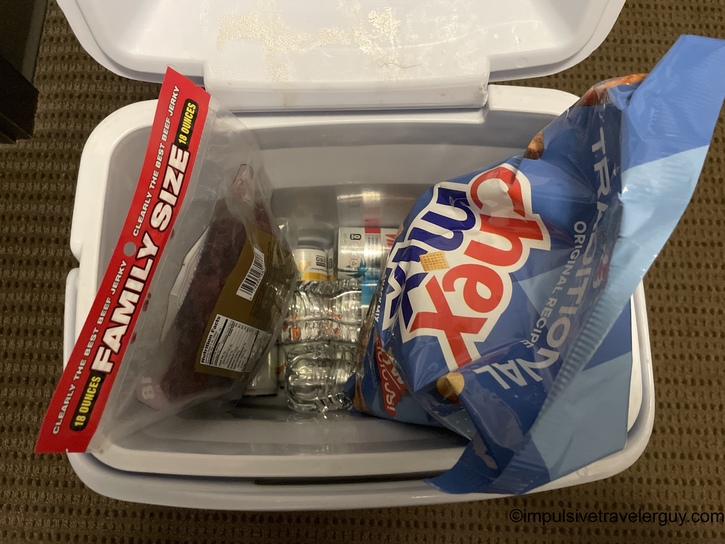 Overhead view of white storage container with snacks including family-size beef jerky package, Auntie Anne's pretzel bag, water bottles, and other items