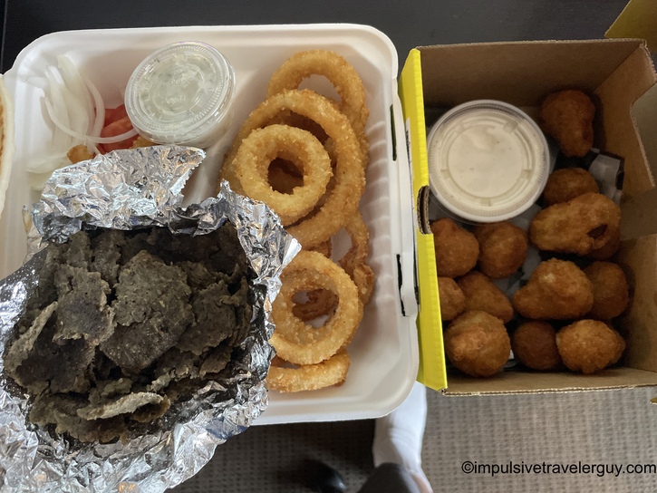 Takeout meal spread showing foil-wrapped gyro or sandwich with onion rings and dipping sauce on white tray, alongside box of breaded cheese curds with ranch dressing