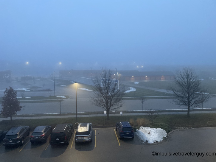 Early morning foggy view from hotel window of empty parking lot with five parked vehicles, bare trees, street lights, and low visibility conditions