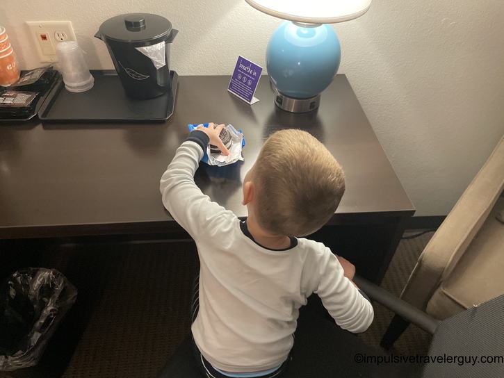 Young child in white long-sleeve shirt reaching for wrapped sandwich at hotel room desk with coffee maker and blue lamp visible