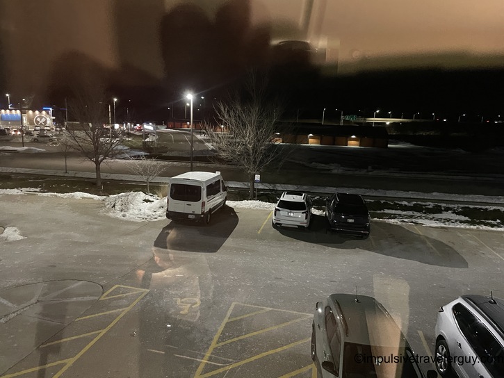 Night view of snow-covered hotel parking lot with white transit van and SUVs, commercial buildings visible in background