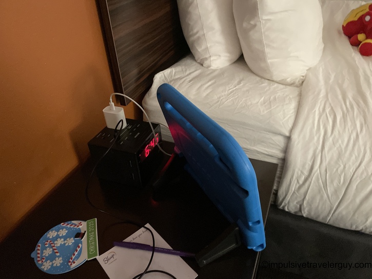 Blue Leapfrog electronic learning tablet propped against pillows on hotel bed with charging station and clock visible on nightstand