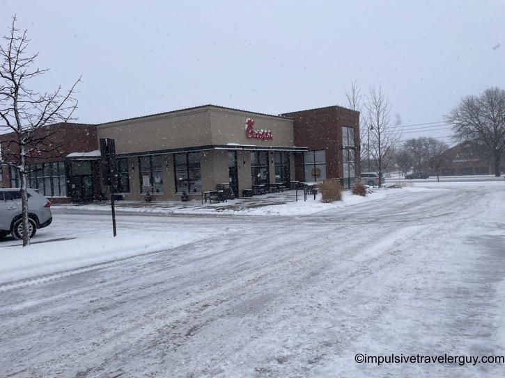 A Chick-fil-A restaurant exterior on a snowy winter day, with snow-covered parking lot and light snowfall visible. The building features the brand's signature modern architecture with large windows and outdoor seating area.