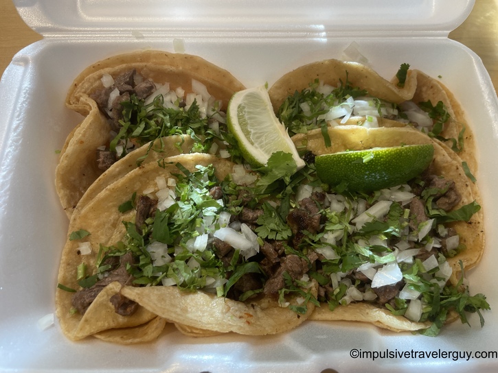Three authentic street-style carne asada tacos on corn tortillas topped with fresh cilantro and diced white onion, served in a white styrofoam container with lime wedges.