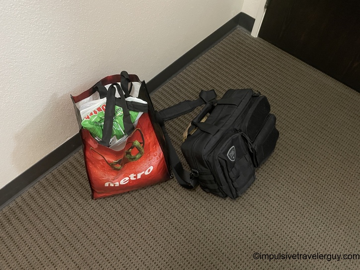 A red Metro grocery store reusable bag and a black tactical-style backpack sitting on a hotel hallway floor against gray carpet and white baseboard.
