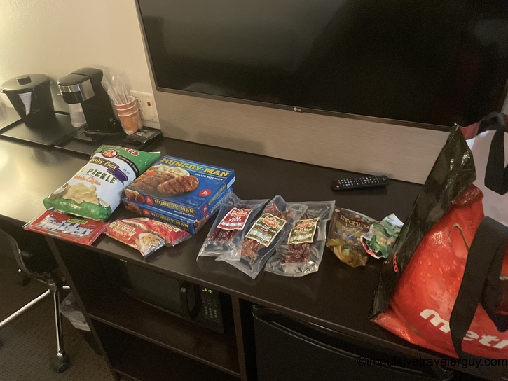 Hotel room desk displaying travel provisions including Hungry-Man frozen dinners, various packages of beef jerky and meat snacks, pickle-flavored chips, and a Metro grocery bag.