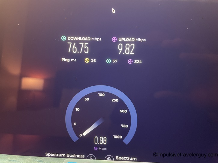 Computer monitor displaying Ookla Speedtest results showing significantly faster hotel WiFi speeds on Spectrum network. Download speed is 76.75 Mbps, upload is 9.82 Mbps, with a speedometer gauge interface visible.
