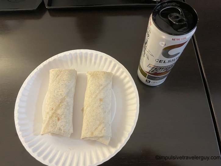 Simple hotel breakfast of two flour tortilla breakfast burritos on a white paper plate, served alongside a can of Celsius energy drink in the "Retro Vibe" flavor, on a dark surface.