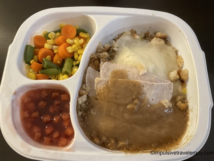 Cooked Hungry-Man turkey frozen dinner in its divided white tray, featuring sliced turkey breast with gravy over stuffing and mashed potatoes, a side of mixed vegetables with corn, carrots, and green beans, and apple cranberry compote.