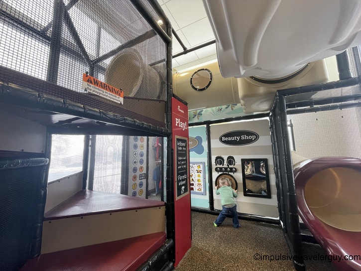 A young child playing in an indoor Chick-fil-A playground, standing at a pretend "Beauty Shop" play station. The play area features climbing structures, slides, and safety netting.