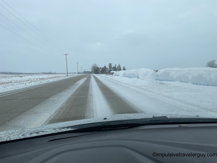 View through a car windshield of a snow-covered rural highway in winter conditions, with flat farmland on both sides and high snow banks along the road edges under an overcast gray sky.