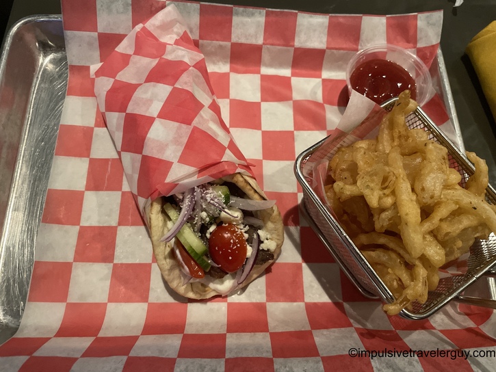 A Greek gyro wrapped in pita bread topped with tomato, cucumber, red onion, and feta cheese, served on red-and-white checkered paper with a side of crispy onion strings and ketchup.