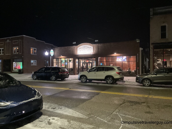 Nighttime street view of "The Garage" restaurant with its illuminated sign, located in what appears to be a small downtown area. Snow is visible on the ground and several vehicles are parked along the street.
