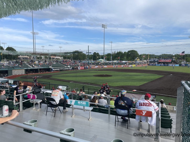 View from the covered grandstand behind the first-base dugout at a Madison Mallards game at Warner Park, with players warming up on the field, protective netting overhead, and YMCA-branded seats in the foreground under a partly cloudy sky