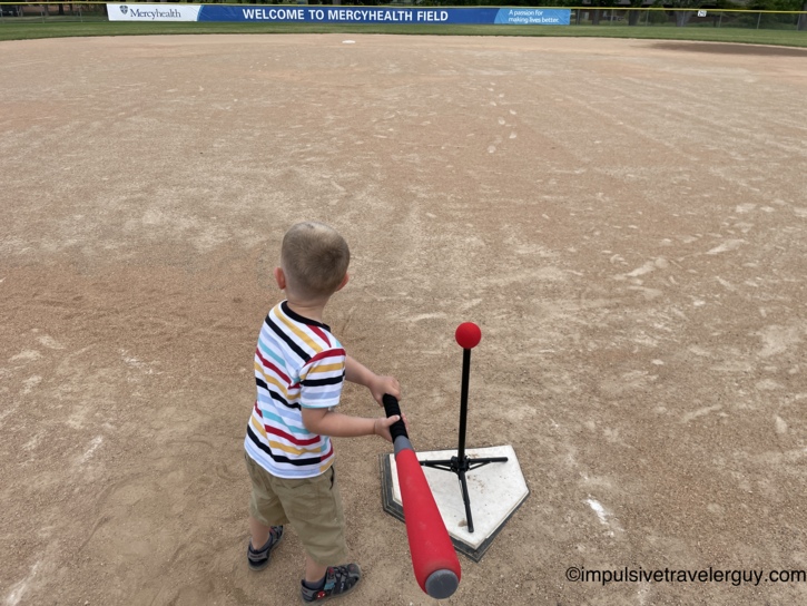 Young child in a striped shirt holding a bat at home plate on an empty dirt ballfield, facing a batting tee with a red ball, beneath a Welcome to Mercyhealth Field outfield banner