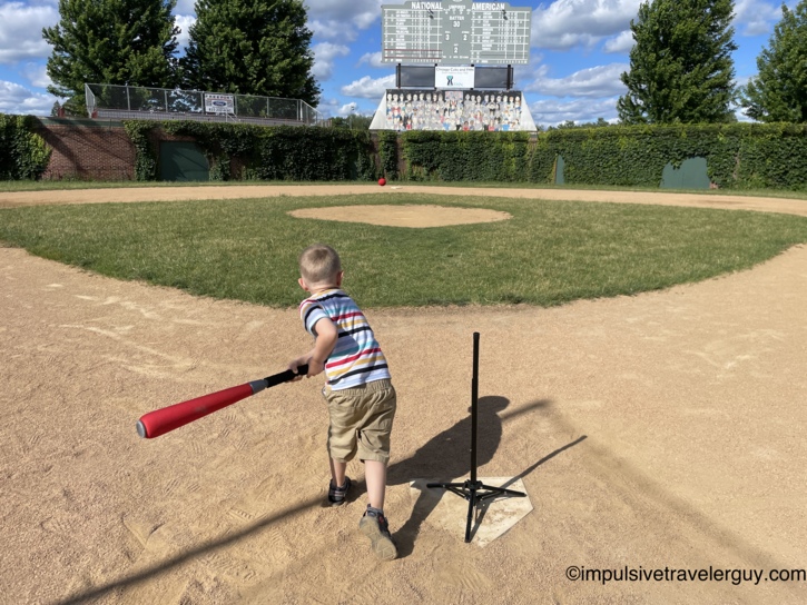 Young child swinging a bat at a batting tee on the field at Little Cubs Field in Freeport, Illinois, with the red ball in the air, an ivy-covered outfield wall, and a replica Wrigley Field scoreboard in the background