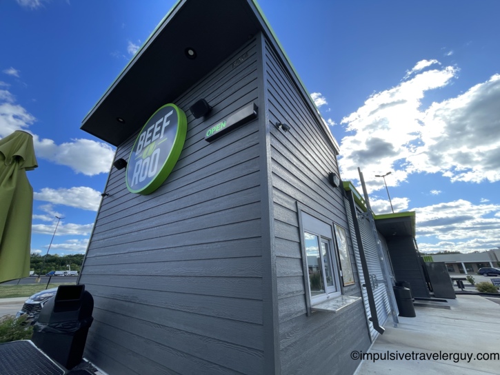 Exterior corner view of a Beef-a-Roo fast-food restaurant with its green circular logo and lit OPEN sign against a bright blue sky