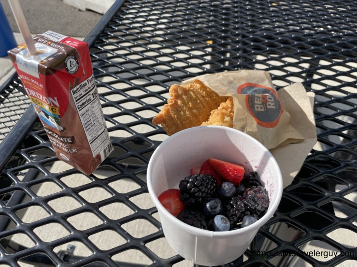 Beef-a-Roo kids meal on a black metal patio table with chicken tenders in a branded bag, a Horizon Organic chocolate milk box, and a cup of mixed fresh strawberries, blackberries, and blueberries