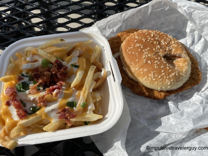 Loaded fries topped with cheese sauce, bacon, green onion, and ranch beside a breaded tenderloin sandwich on a sesame-seed bun, on a black patio table at Beef-a-Roo