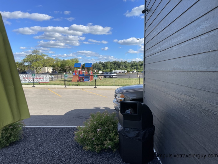 View from a Beef-a-Roo patio toward a parking lot and a fenced dog park with wooden play structures and a dog, with a Shoe Sensation banner on the fence under a blue sky