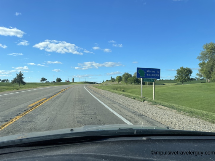 View through a car windshield of a rural highway approaching a green Welcome to Wisconsin, Green Co road sign, with farmland and a barn in the distance under a clear blue sky
