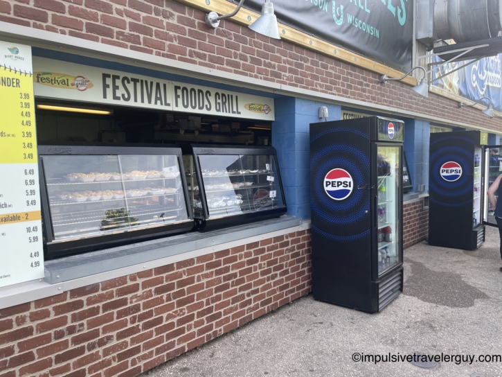 Festival Foods Grill concession stand at Warner Park showing a yellow menu board with prices, a heated food display case of brats and sandwiches, and two illuminated Pepsi refrigerators against a brick and blue wall