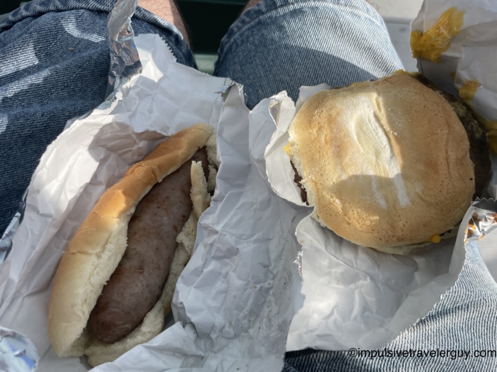 Close-up of a bratwurst in a bun and a cheeseburger held in foil wrappers on a lap in blue jeans, ballpark concession food at a Madison Mallards game