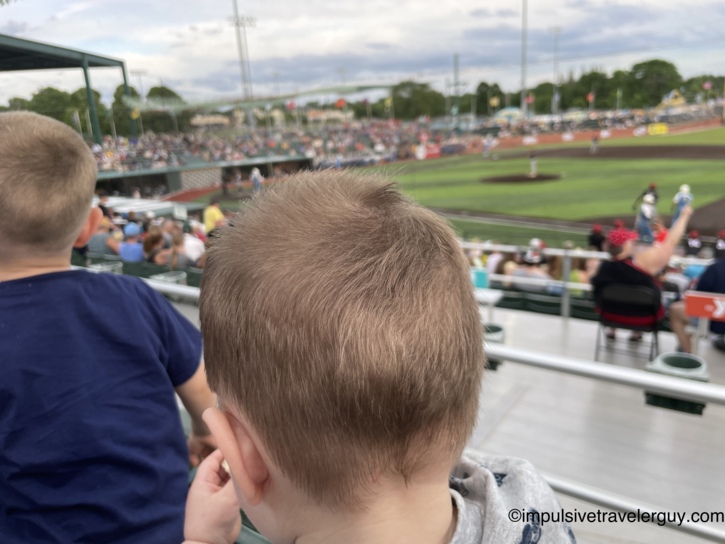 View over the back of a young child's head watching a Madison Mallards game from the grandstand, with a crowded stadium, green infield, and a mascot visible in the blurred background