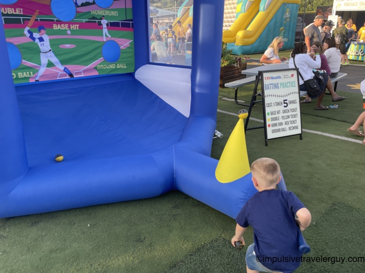 Young child holding a bat at an inflatable batting-practice carnival game in the kids zone at a Madison Mallards game, with a UW Health Sports Medicine Batting Practice sign and bounce houses nearby