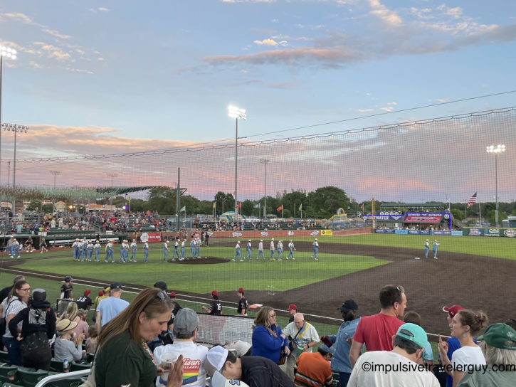 Wide view of the baseball field at sunset during a Madison Mallards game, with players and staff gathered on the infield, a pink and blue sky overhead, and fans watching from the grandstand