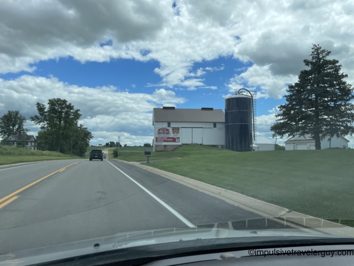 View through a car windshield of a rural road passing a white barn painted with a billboard for Little Cubs Field, a Wrigley Field replica in Freeport, Illinois, beside a dark silo under a cloudy sky