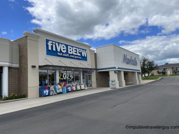 Exterior of a strip-mall retail center showing Five Below and Marshalls storefronts, with 2026 foil balloons in the Five Below window and pool floats on display, under a partly cloudy sky