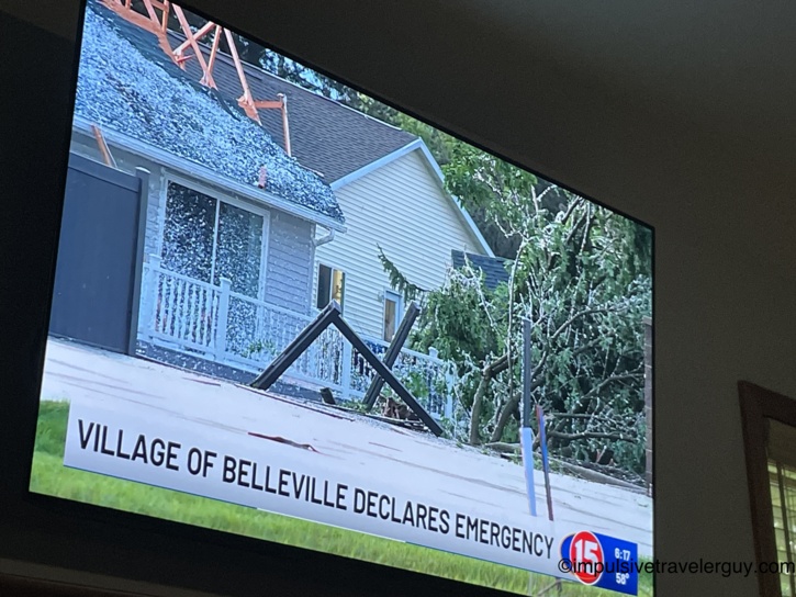 Television screen showing Channel 15 news broadcast with the chyron 'Village of Belleville Declares Emergency,' showing a damaged house with exposed roof trusses, a downed tree, and bent porch railing from storm damage.