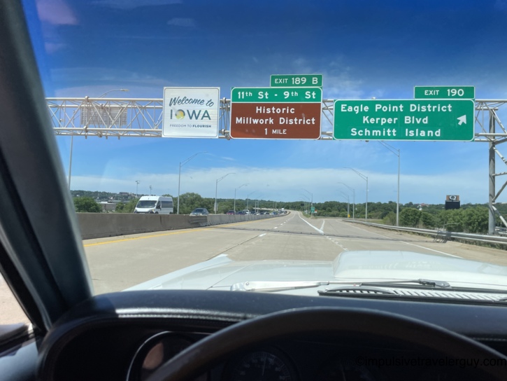 Crossing the Mississippi River bridge into Iowa in the Cougar, passing the 'Welcome to Iowa — Freedom to Flourish' sign alongside green overhead signs for the Historic Millwork District, Eagle Point District, Kerper Blvd, and Schmitt Island in Dubuque.