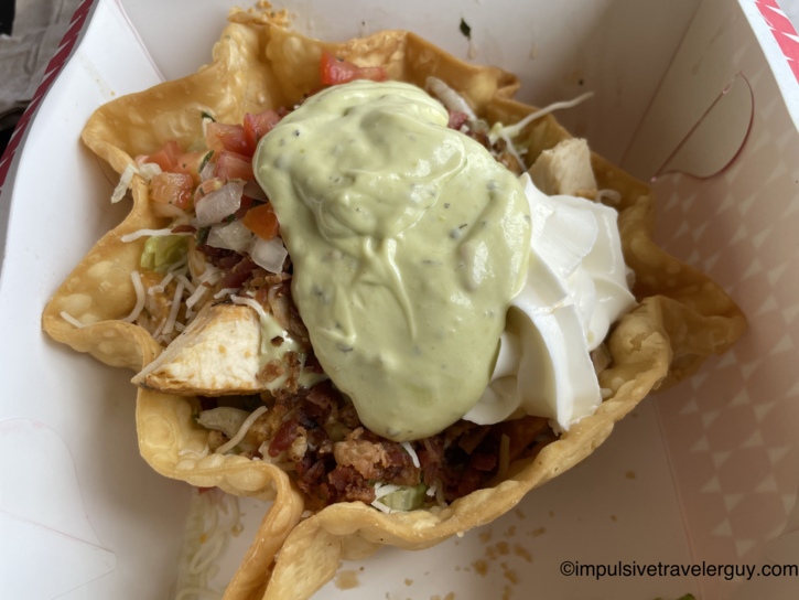 A Taco John's taco salad in a crispy tortilla bowl loaded with grilled chicken, bacon bits, shredded cheese, pico de gallo, sour cream, and a generous dollop of avocado crema.