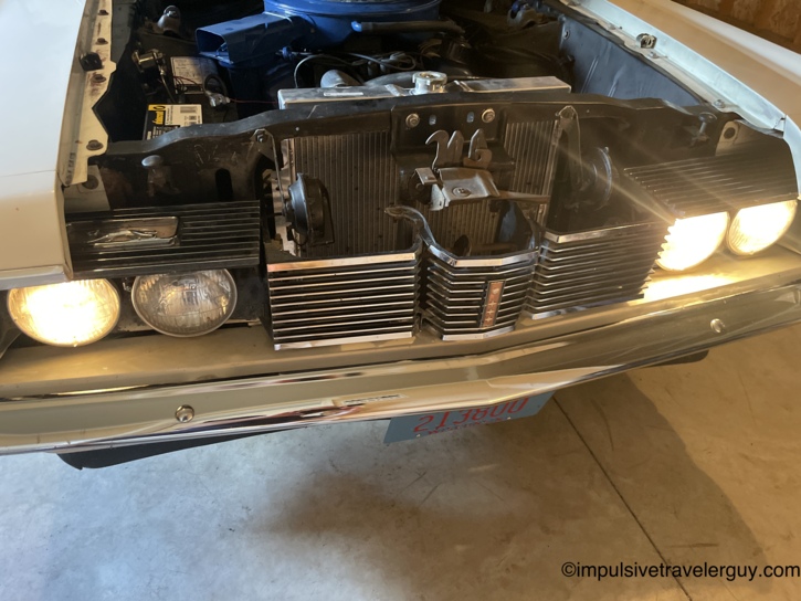 Front of the Cougar in the garage with the hood up and headlights on, shot from a low angle looking up past the chrome bumper and grille — the driver-side inner headlight noticeably dimmer than the others.