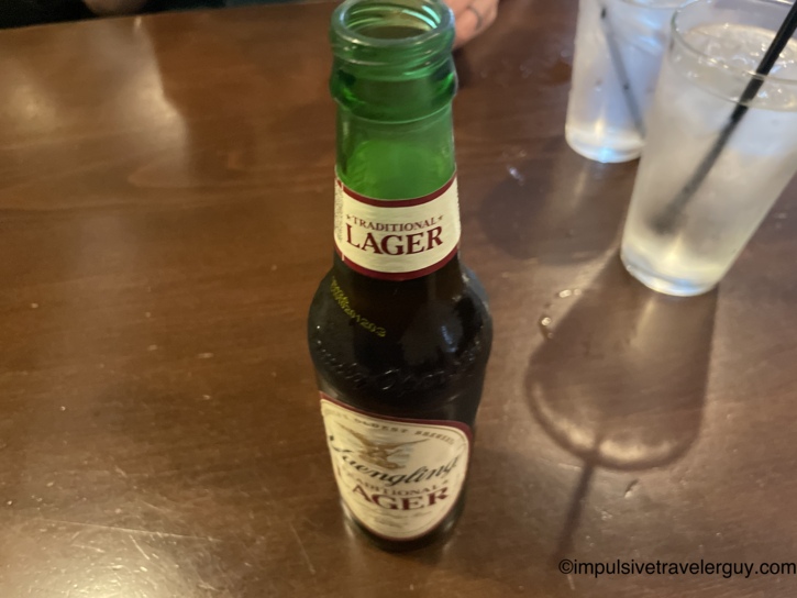 Yuengling Traditional Lager bottle on a dark wooden bar table with water glasses in the background.