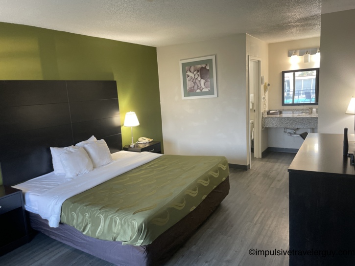 Budget motel room with king bed, green accent wall, dark headboard, patterned bedspread, and open vanity area with granite countertop.