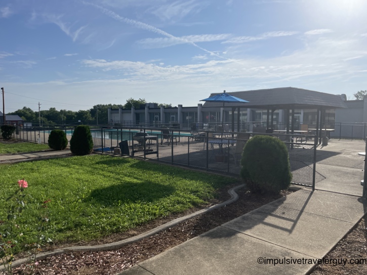 Motel pool and patio area in morning sunlight with fenced deck, blue umbrella, covered pavilion, and manicured lawn.