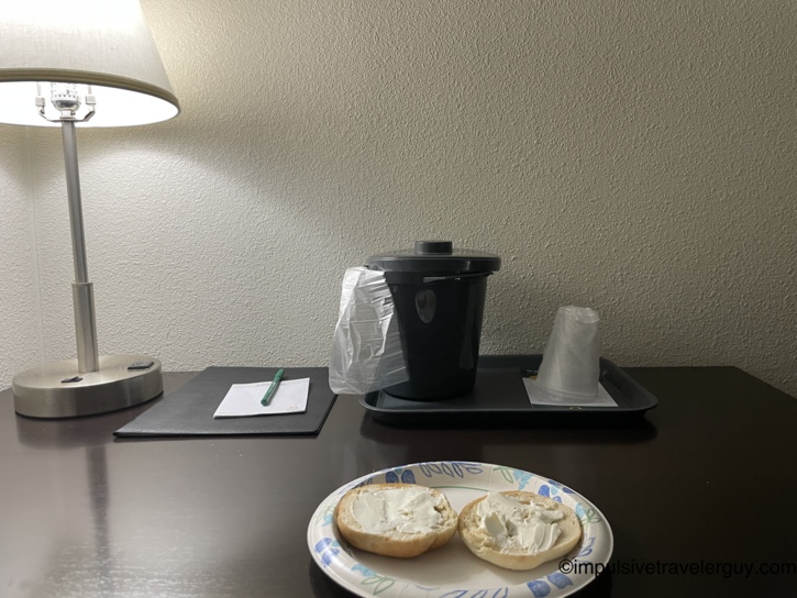 Cream cheese bagel on a paper plate on the motel room desk next to a desk lamp, notepad, and ice bucket.