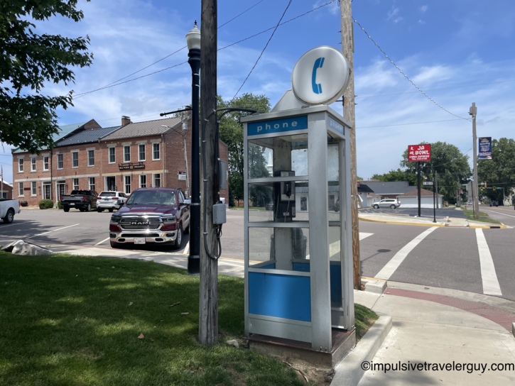 Vintage glass-panel phone booth with blue accents and a round phone symbol on top, standing on a small-town street corner with historic brick buildings and a bowling alley sign in the background.